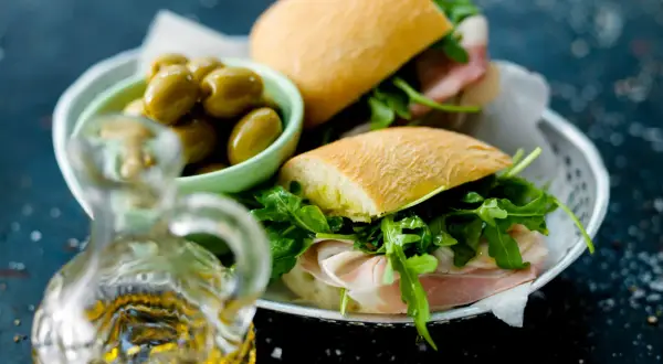 Två halvor av en ciabatta macka med prosciutto och ruccola, serverade med gröna oliver och en karaff olivolja.