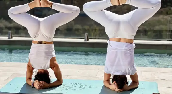 Dos mujeres en postura de Sirsasana, con las piernas abiertas y juntas en **hasta bandha**, practican yoga junto a una piscina.