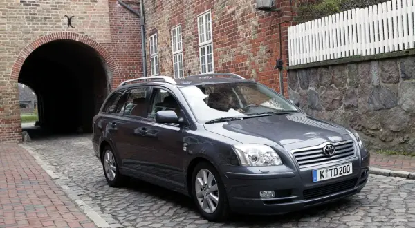 Szary Toyota Avensis T25 2.0 D4D na brukowanej uliczce, obok ceglanego budynku z łukiem.