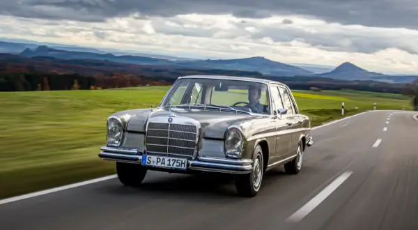 En klassisk Mercedes 60-tal glider fram på en slingrande landsväg med böljande gröna fält och dramatiska berg i bakgrunden.