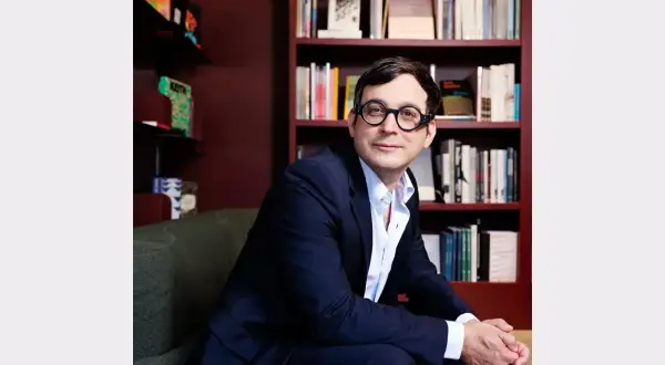 A man in a suit and glasses sits in front of bookshelves, perhaps at the David Lewis Gallery, looking thoughtfully to the side.
