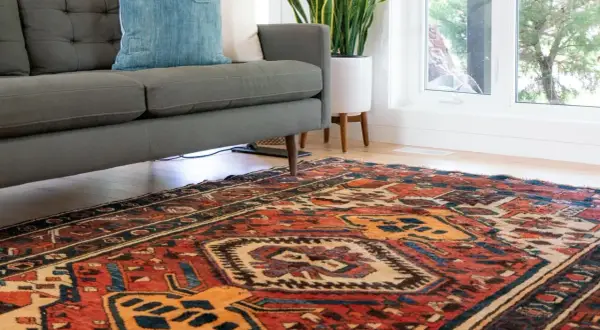 Ein detailreicher perserteppich mit roten und blauen Mustern liegt vor einem grauen Sofa. Tipps zum perserteppich reinigen.