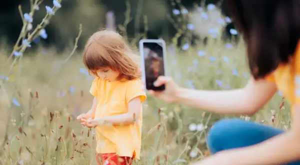Dziecko w żółtej koszulce zbiera kwiatki na łące, a mama dokumentuje chwilę telefonem, praktykując sharenting.