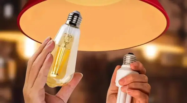 Une ampoule connectée orange, au design vintage, est tenue à côté d'une ampoule fluocompacte, sous un abat-jour rouge.
