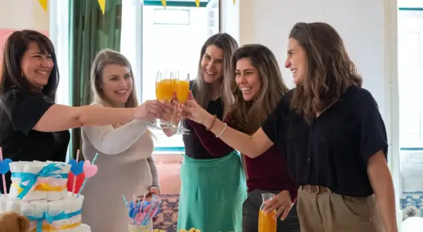Przyjęcie baby shower: kobiety wznoszą toasty, przyszła mama w beżowym swetrze. Idealna stylizacja na baby shower to coś wygodnego i eleganckiego.