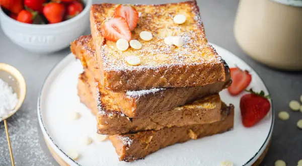 Gestapelte French Toast Scheiben, bestäubt mit Puderzucker und garniert mit Erdbeeren und weißen Schokoladenchips.