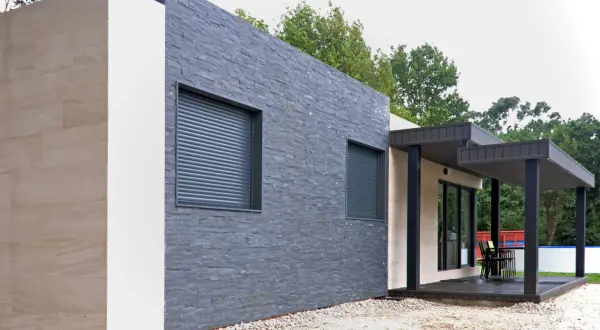 Muros exteriores de casas modernas con texturas de piedra y acabados claros. Ventanas con persianas enrollables y un porche con mobiliario.