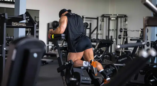 Man utför stående lårcurl på ett gym. Han har en svart tröja och shorts, och tränar sina baksida lår.