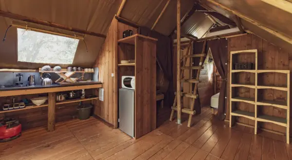 Intérieur d'une cabane en bois avec coin cuisine, réfrigérateur, étagères et échelle menant à un lit. Un havre de paix en Haute-Loire.