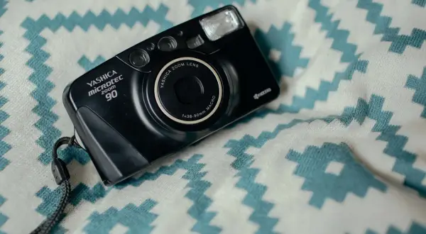 Czarny aparat Yashica Microtec Zoom 90 z paskiem, leżący na białym materiale z turkusowymi zygzakami. Wygląda jak z lat 90.