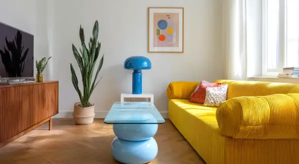 Jasny salon retro z żółtą kanapą, niebieską lampą i stolikiem kawowym. Drewniana komoda i duża roślina dopełniają przytulną atmosferę.