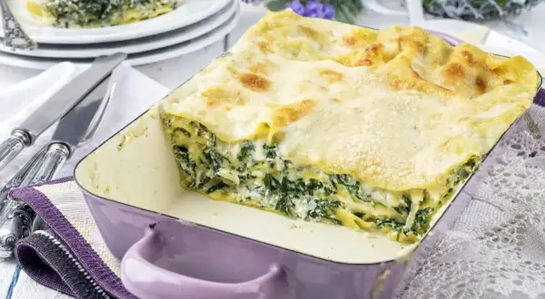 Pyszne lasagne ze szpinakiem w fioletowym naczyniu, gotowe do podania. Obok sztućce i dekoracyjne kwiaty.