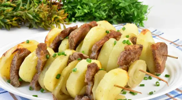 Karkówka pieczona w plastrach z cebulą i ziemniakami na patyczkach, posypana szczypiorkiem.