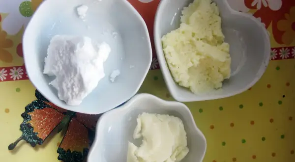 Trzy białe miseczki z białymi i żółtymi składnikami do domowych kosmetyków. W jednej proszek, w drugiej grudki, a w trzeciej gładka masa. Obok motyl. To składniki do zrobienia własnego masła shea.