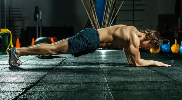 Mann in Unterarmstützposition, um zu zeigen, wie oft bauchmuskeln trainieren. Im Hintergrund sind Kettlebells und Trainingsgeräte.