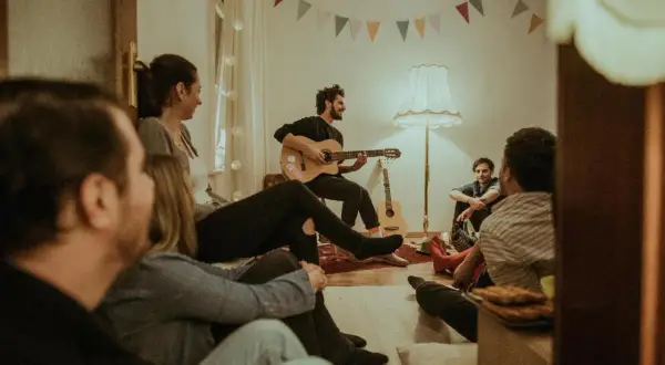 Freunde versammeln sich für einen gemütlichen Abend mit Gitarrenmusik und **Lieder zum Mitsingen**. Bunte Wimpelketten schmücken die Wand.