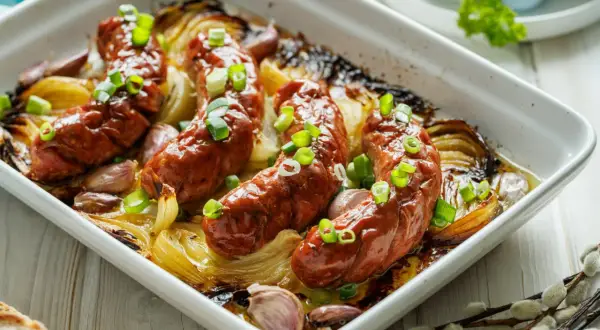 Pieczona biała kiełbasa surowa z cebulą i czosnkiem w naczyniu żaroodpornym, posypana szczypiorkiem.