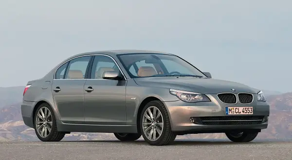 Szary sedan BMW 5 Series (E60) z jasnym wnętrzem stoi na pustej drodze, z górami w tle.
