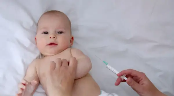Małe dziecko leży na białym tle, a ręka pielęgniarki przygotowuje szczepienia noworodka w szpitalu.