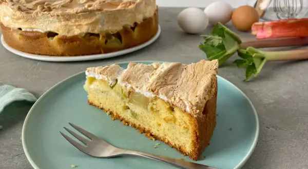 Ein saftiges Stück Rhabarber Baiser Kuchen mit einer luftigen Baiserhaube liegt auf einem blauen Teller. Im Hintergrund ein ganzer Kuchen und frischer Rhabarber.