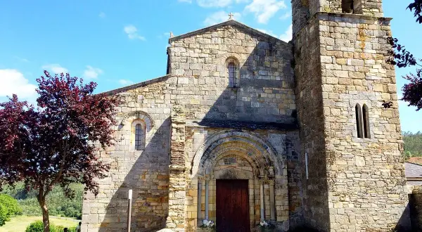 La catedral de Foz, con su torre románica y fachada de piedra, se alza bajo un cielo azul con nubes. Un árbol de hojas rojizas añade color.