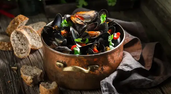 Un grand pot en cuivre rempli de moules ouvertes avant cuisson, garnies de persil et de piment. Du pain rustique est disposé sur la table en bois.