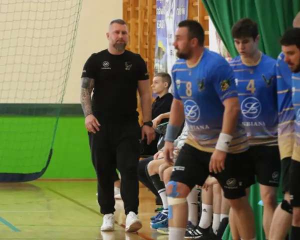 Maciej Reichel, trener, obserwuje mecz piłki ręcznej. Zawodnicy w niebieskich koszulkach czekają na swoją kolej.