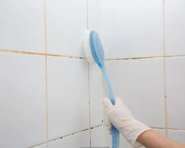 Mano con guanto bianco usa spazzola blu per pulire le fughe delle piastrelle bianche, mostrando come togliere la muffa.
