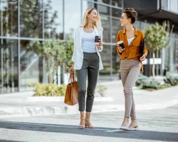 Dos mujeres con looks para ir a trabajar, tomando café y charlando, caminan frente a un edificio moderno.