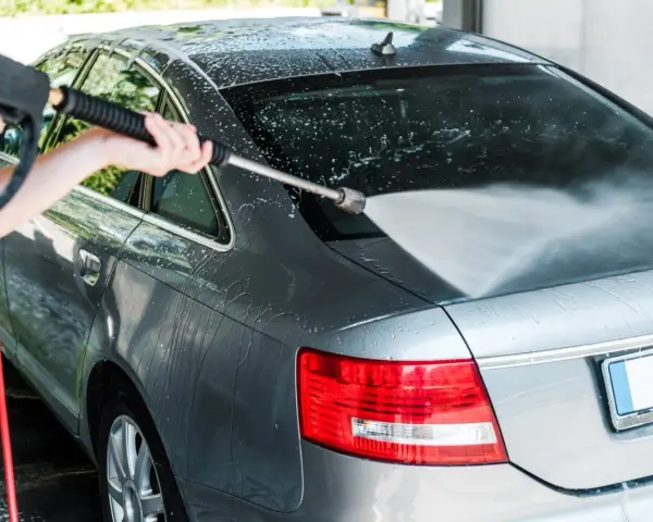 A person demonstrates how to power wash a car, spraying water onto the rear of a grey sedan.