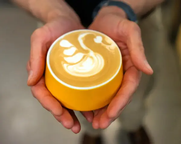Dłonie trzymają żółtą filiżankę z kawą, na której widnieje wzór łabędzia. Zastanawiasz się, ile kofeiny ma espresso?