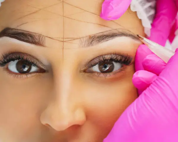 Nahaufnahme einer Frau, deren Augenbrauen mit Linien markiert werden, während eine Person in pinken Handschuhen mit einem Stift arbeitet.