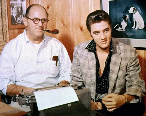 Tom Parker und Elvis Presley sitzen nebeneinander. Parker, mit einer Pfeife im Mund, tippt auf einer Schreibmaschine.