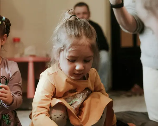 Skupiona dziewczynka w pomarańczowej bluzce, jak bawi się dziecko z autyzmem, z uwagą przygląda się swoim dłoniom.
