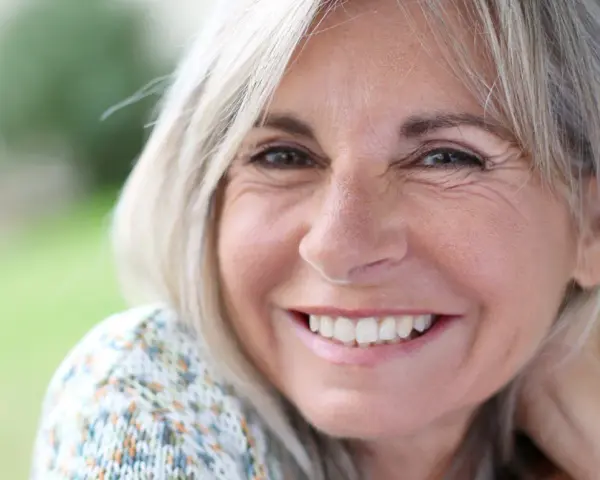 Mujer sonriente con cabello rubio y arrugas normales a los 50 años, disfrutando de un día al aire libre.
