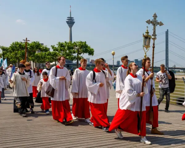 Jugendliche in Soutanen und Alba nehmen an einer Prozession teil, die an christliche Feiertage erinnert. Sie tragen Kreuze und Fahnen.