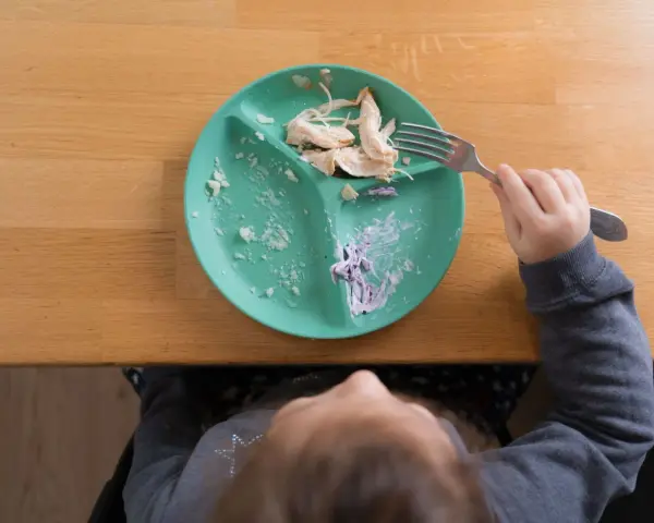 Dziecko je kurczaka z talerza babyono z przyssawką. Resztki jedzenia na talerzu i widoczna rączka z widelcem.