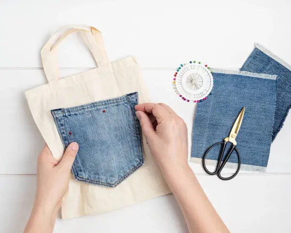 Mains épinglant une poche extérieure en jean sur un sac en toile. Ciseaux et chutes de tissu à proximité.