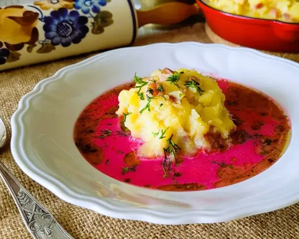 Pyszny barszcz czerwony zabielany, przepis babci. Na talerzu purée ziemniaczane z dodatkiem boczku i koperku, zanurzone w intensywnie różowym barszczu.
