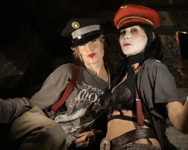 Deux femmes aux maquillages théâtraux portent des casquettes et des bretelles, l'une tenant un téléphone.