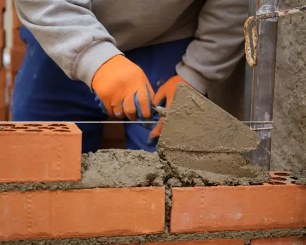Budowa murowanego prostego grilla z cegły. Ręce w pomarańczowych rękawiczkach nakładają zaprawę kielnią.