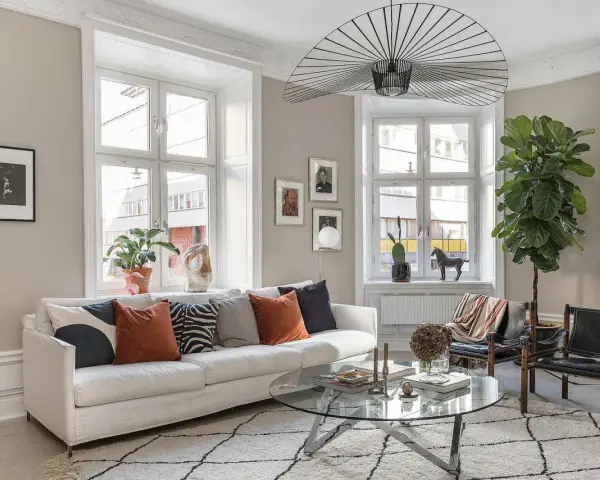 Vardagsrummet är ljust med en vit soffa, kuddar i olika färger och mönster, ett runt soffbord och en stor taklampa. En stor grön växt bidrar till trivseln.