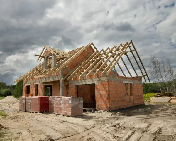 Budowa domu 100m2 w toku. Ściany z cegły, drewniany szkielet dachu, materiały budowlane na placu.