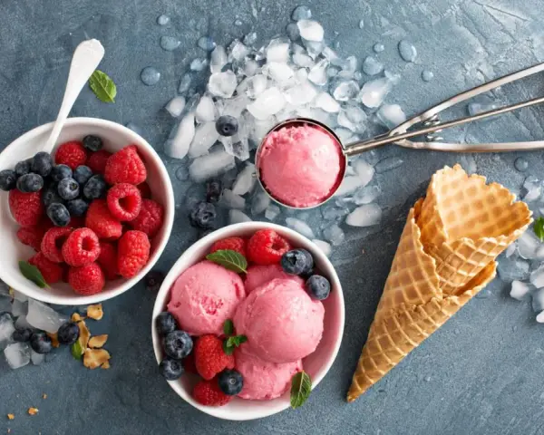 Himbeeren und Blaubeeren neben Schalen mit leckerem Milcheis selber machen. Eiswaffeln und ein Eisportionierer liegen bereit.