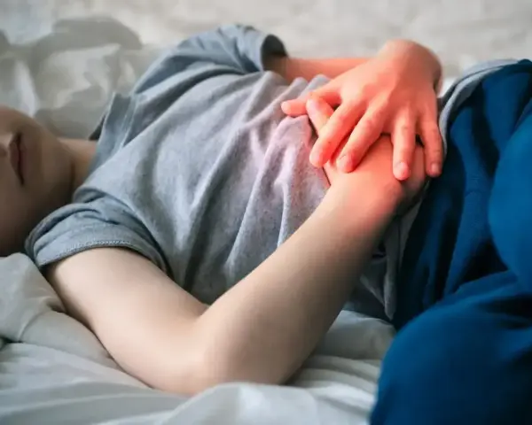 Dziecko leży w łóżku, trzymając się za brzuch. Widać zaczerwienienie sugerujące ból brzucha u dziecka.