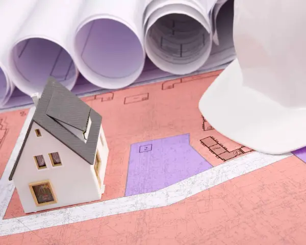 Model domu na planach, biały kask budowlany i zwoje dokumentacji. Rozważanie służebności gruntowej przy planowaniu inwestycji.
