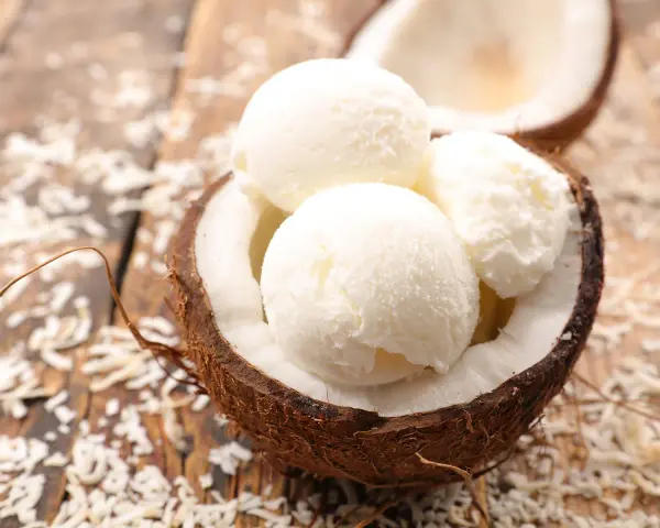 Cremiges Kokosnuss Eis in einer halbierten Kokosnuss, umgeben von Kokosraspeln auf Holz. Ein tropischer Genuss!