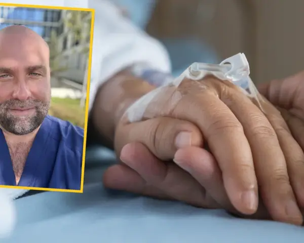 Uśmiechnięty mężczyzna w niebieskim uniformie medycznym podaje słowa otuchy choremu na raka, trzymając go za rękę z wenflonem.