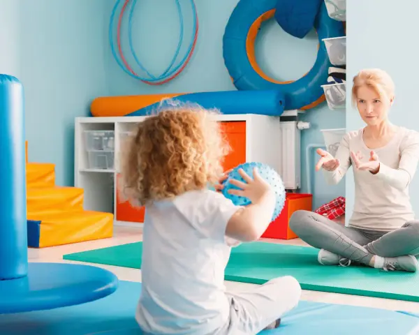 Ergotherapie für Kinder: Eine Therapeutin leitet ein Kind mit lockigem Haar an, das einen blauen Ball hält, in einem bunten Raum voller Spielgeräte.