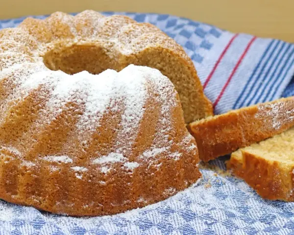 Puszysta babka z cukrem pudrem, obok niej kromki ciasta, na niebieskim ręczniku.